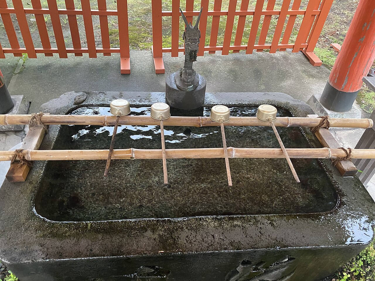 Stone shrine purification basin with bamboo ladles and a bronze dragon spout beside a red fence