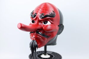 Red tengu mask with a long protruding nose, black eyebrows and a black feathered beard on a display stand