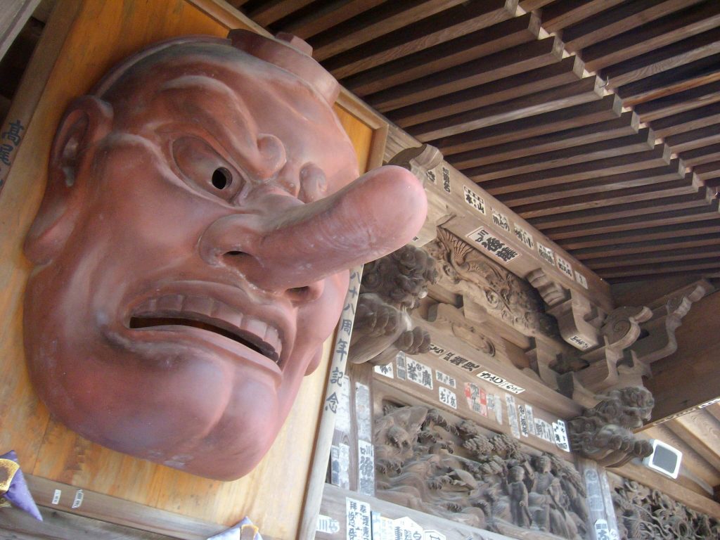Giant carved wooden daitengu face with a long nose and bared teeth mounted under the eaves of a temple hall
