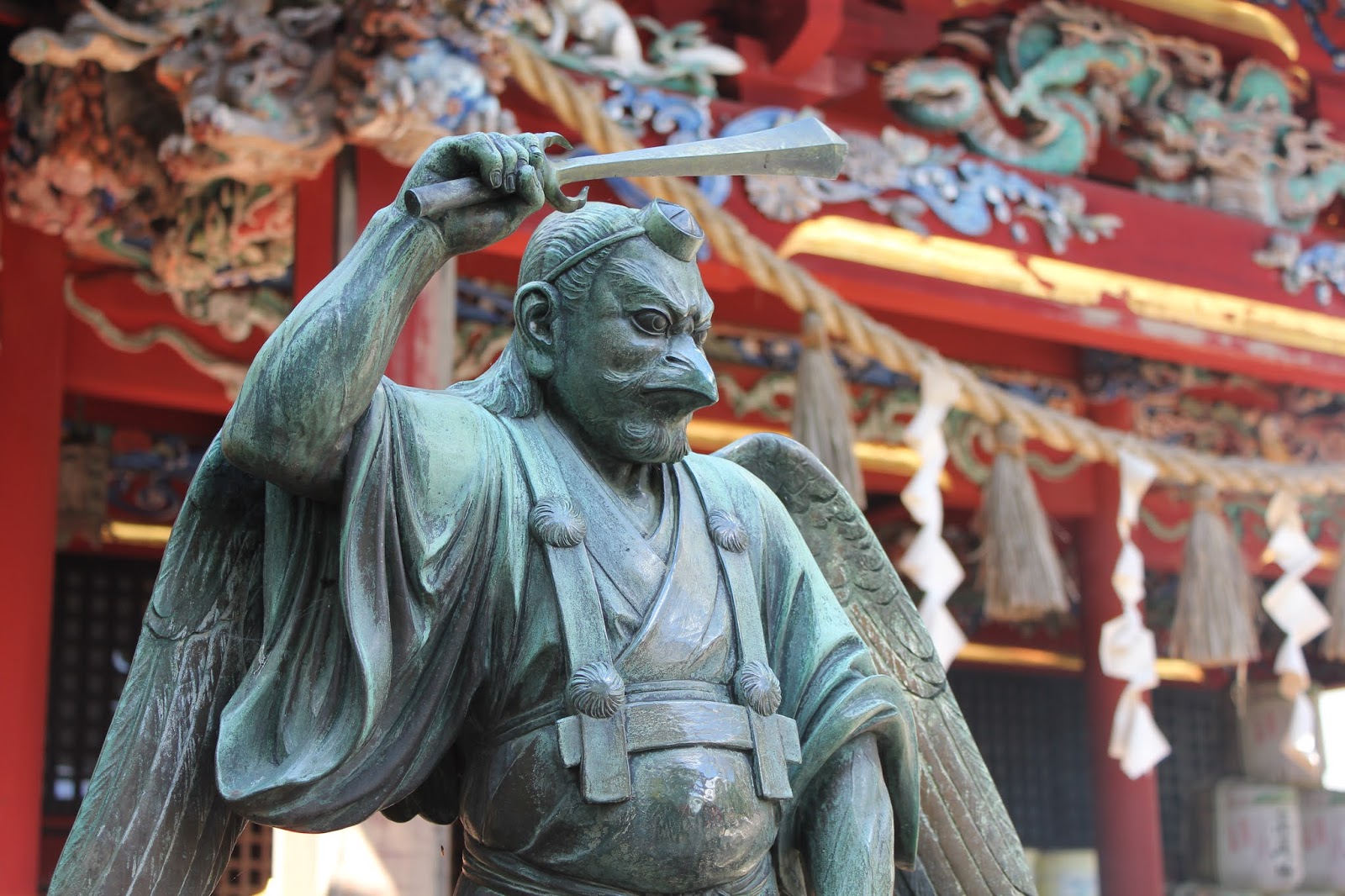 Bronze karasu tengu statue with a beak, folded wings, tokin cap and a raised sword standing before a red temple hall