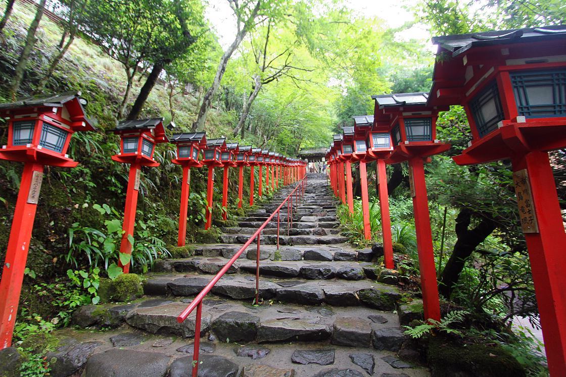 Stone steps lined with red wooden lanterns climbing through dense green forest on the Kurama to Kibune route north of Kyoto