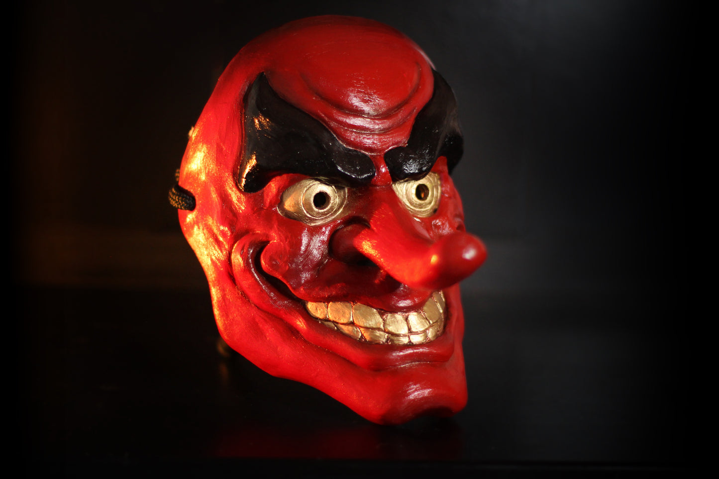 Lacquered red tengu mask with gold teeth, black eyebrows and a jutting nose lit against a dark background