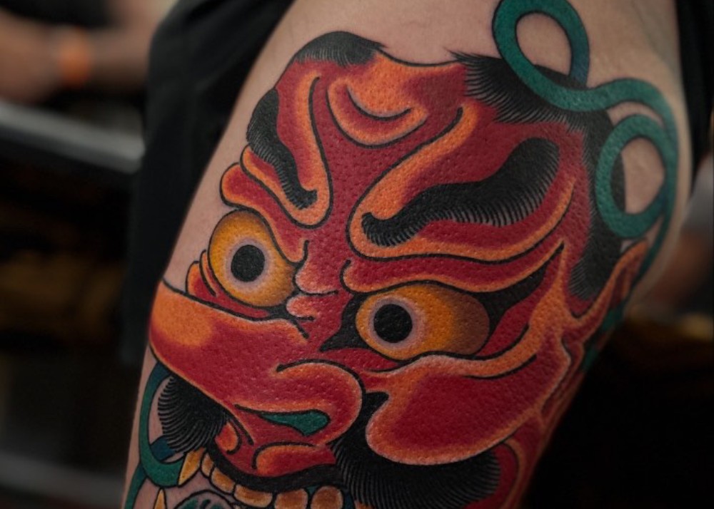 Color tattoo of a red-faced daitengu mask with a long nose on a person's upper arm