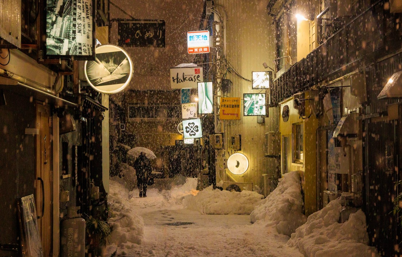 Lone figure with an umbrella walking down a snow filled Japanese alley lit by shop signs at night