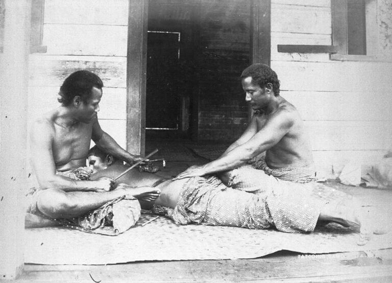Black-and-white photograph of a Samoan man being tattooed by two men seated on a veranda, circa 1895