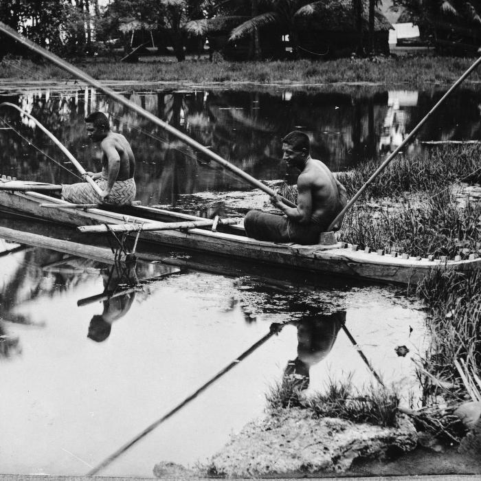 Black-and-white photograph of two shirtless men sitting in a wooden outrigger canoe on still water, holding long fishing poles