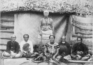 Black-and-white photograph of Mataʻafa Iosefa standing in front of a thatched house, with his wife and four chiefs seated on mats, about 1900