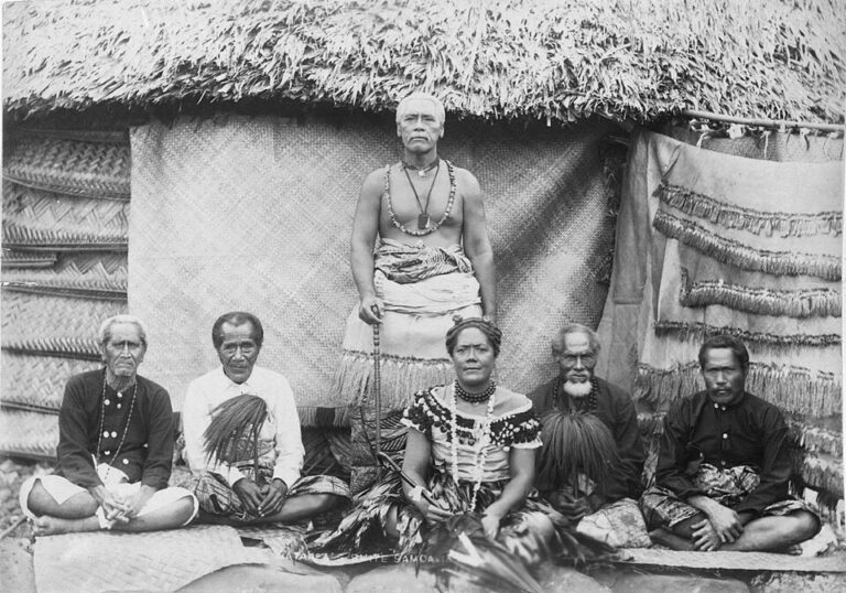 Black-and-white photograph of Mataʻafa Iosefa standing in front of a thatched house, with his wife and four chiefs seated on mats, about 1900