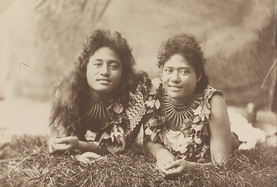 Sepia photograph of two young Samoan women with long hair lying on grass, wearing leaf garlands and tooth necklaces